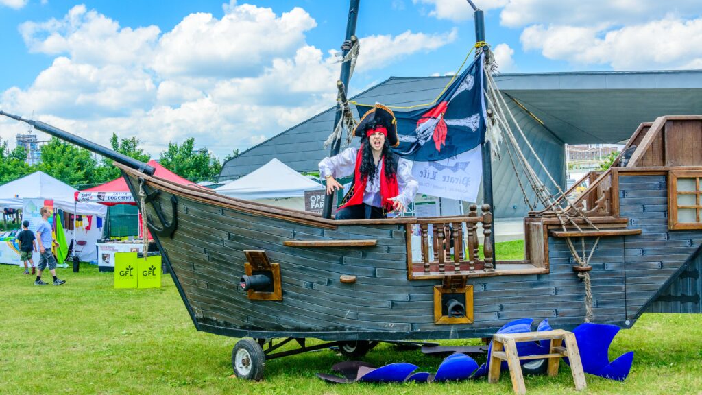 Wooden pirate ship with wheels on grass at a past toronto waterfront festival event. Pirate is seen standing on the deck of the ship. 